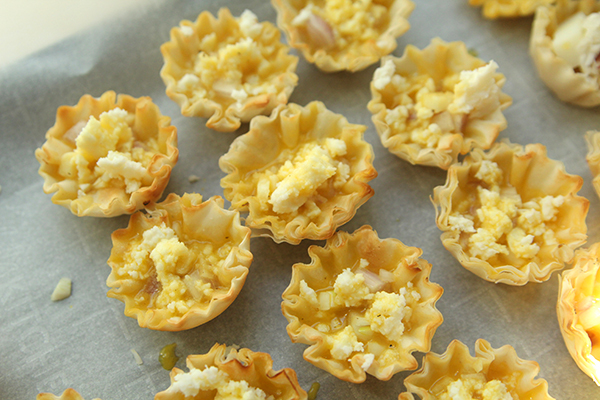 Frilly-edged shells hold liquid egg and crumbled cheese. They are sitting on parchment paper.