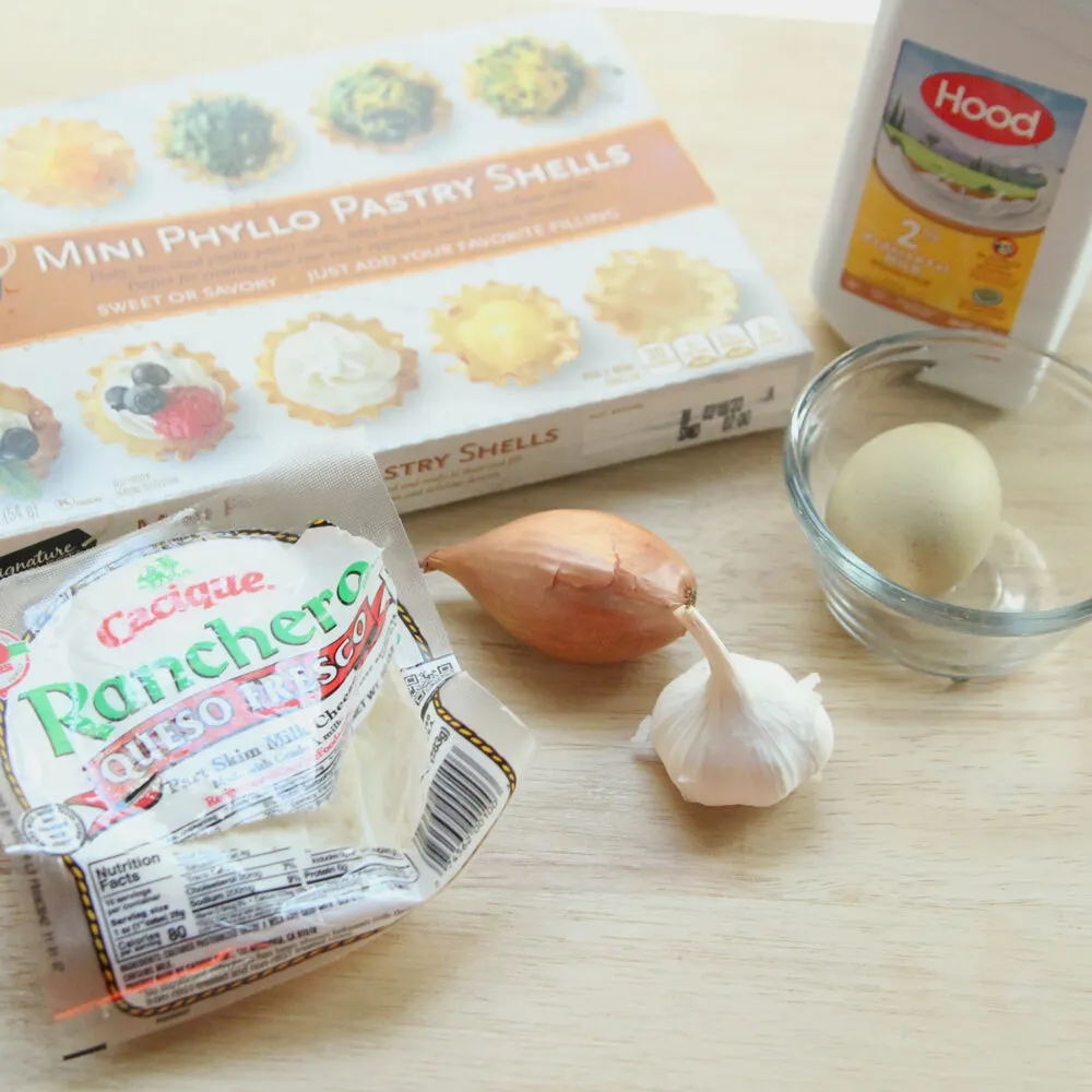 A box of Mini Phyllo Pastry Shells sits on a wooden counter with an open package of queso fresco, a head of garlic, a shallot, an unbroken egg in a bowl and a bottle of milk.