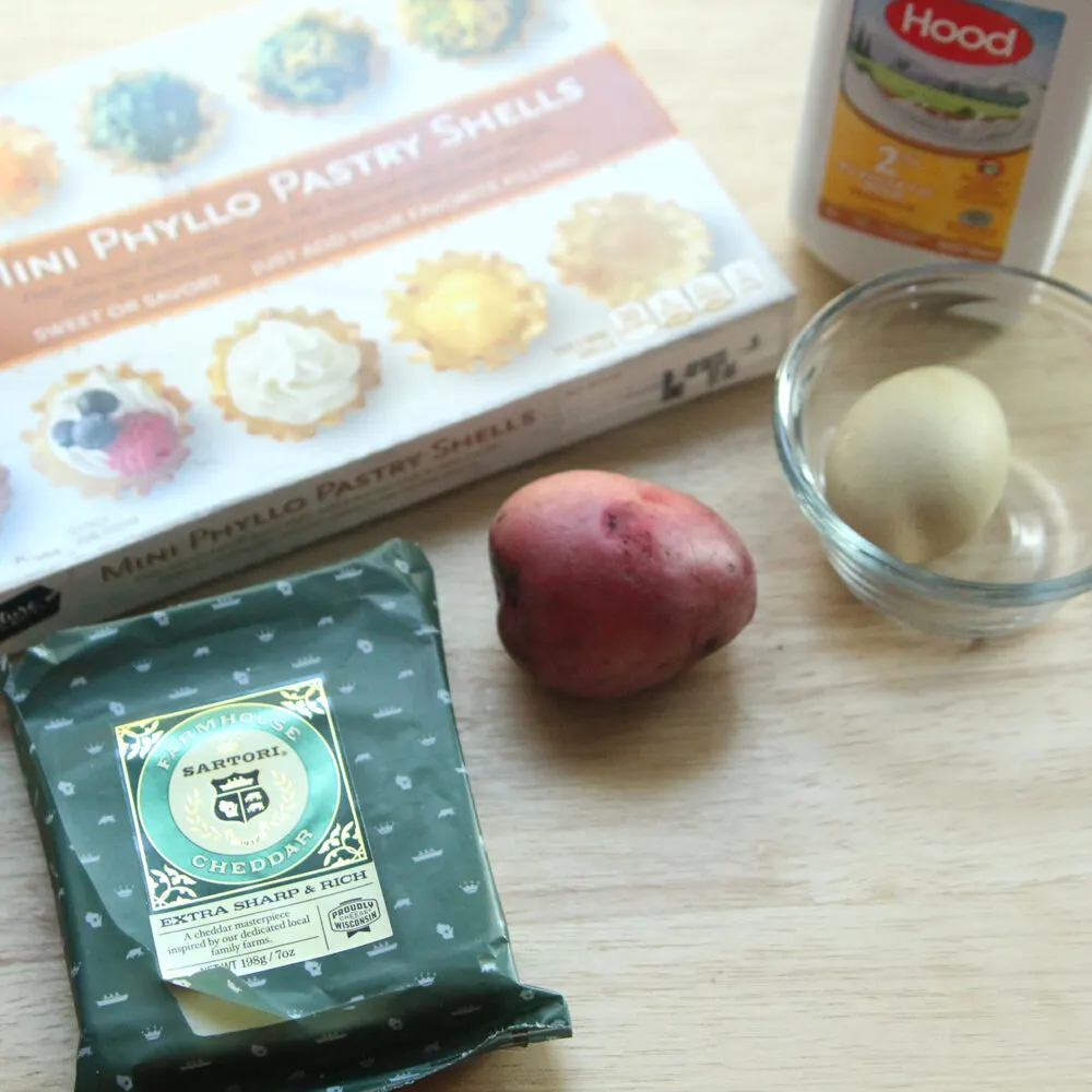 On a wooden surface sits an egg in a glass bowl, a block of cheddar cheese in a green package, a package of Mini Phyllo Pastry Shells, a red potato and milk.