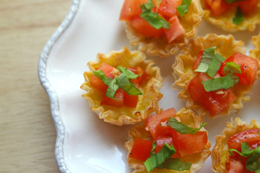 On a white plate on a wooden surface, ruffle-edged light brown fillo shells hold a faintly yellow mixture topped with red tomatoes and green basil.
