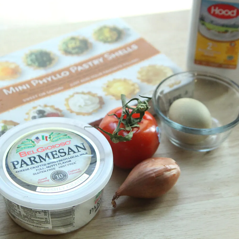 On a wooden surface sits a plastic container of parmesan cheese, a red tomato with a green stem, a brown shallot, a green-shelled egg and a box of mini fillo shells. A bottle of milk sits in the background.