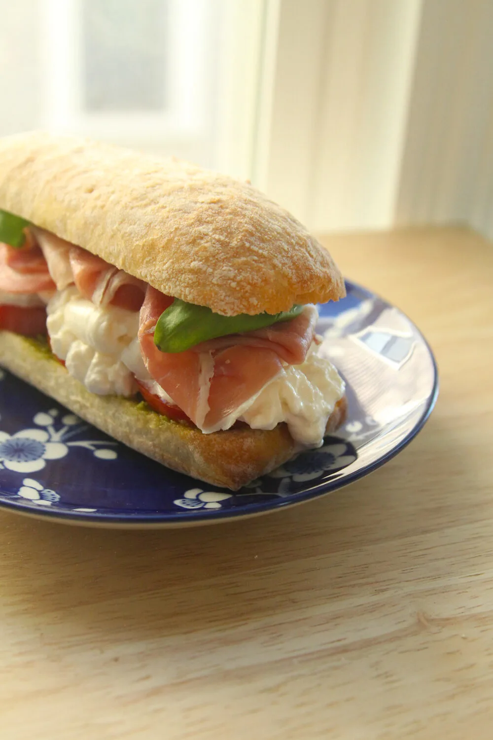 A sandwich on tan ciabatta bread is shown on a blue and white plate sitting on a wooden countertop. It has red tomatoes, white burrata cheese, pink prosciutto and green basil visible.