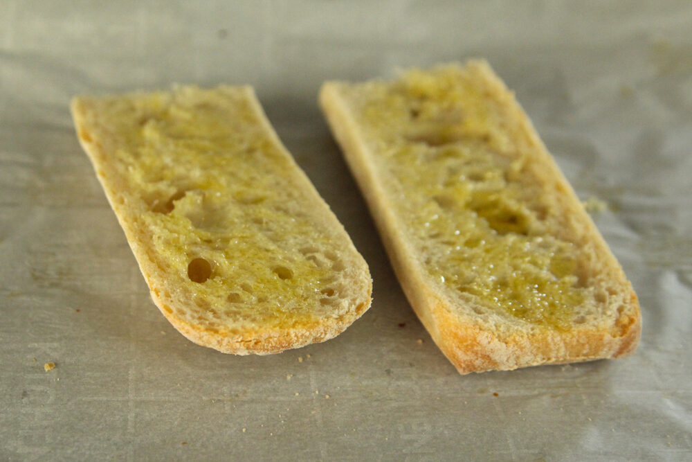 Two halves of a ciabatta loaf sit on a parchment lined baking sheet. The yellow of olive oil that has been brushed on the cut side is visible.
