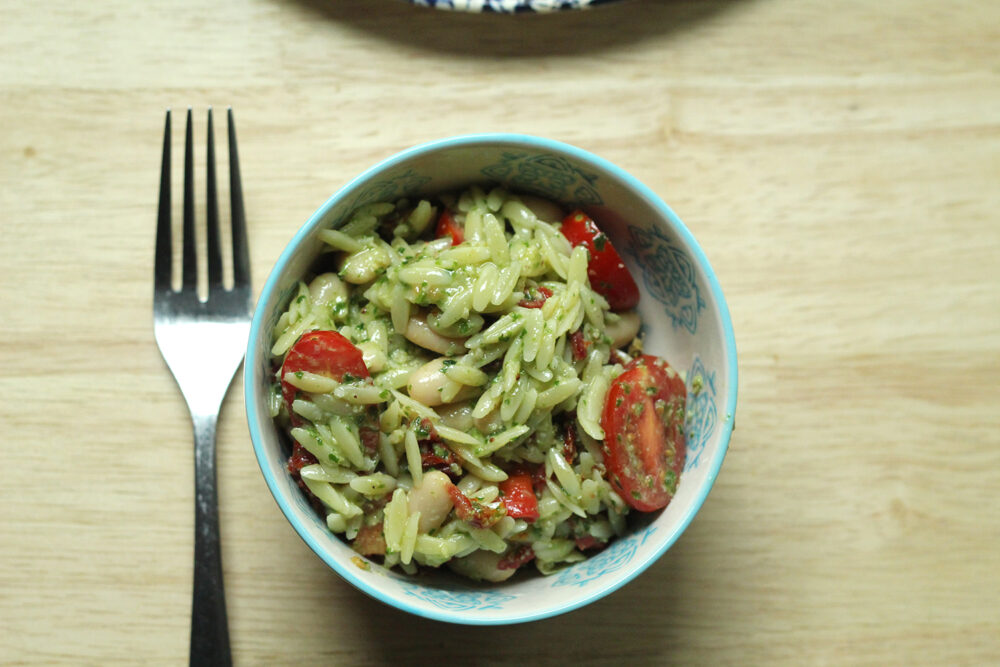 A light blue and white patterned bowl holds oblong orzo, red tomatoes, and creamy colored beans with a green flecked sauce. 