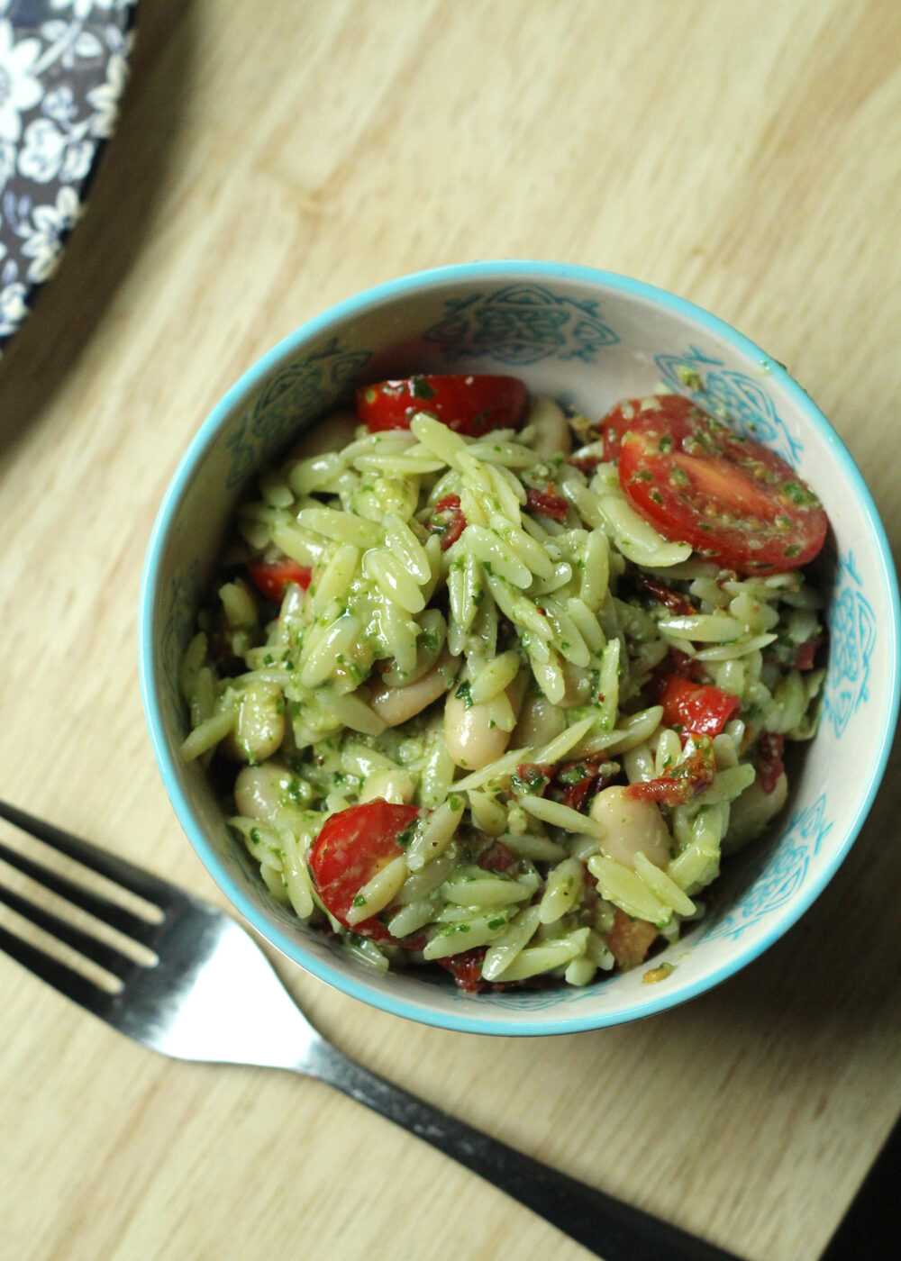 A light blue and white patterned bowl holds an oblong orzo, red tomatoes, and creamy colored beans with a green flecked sauce. 