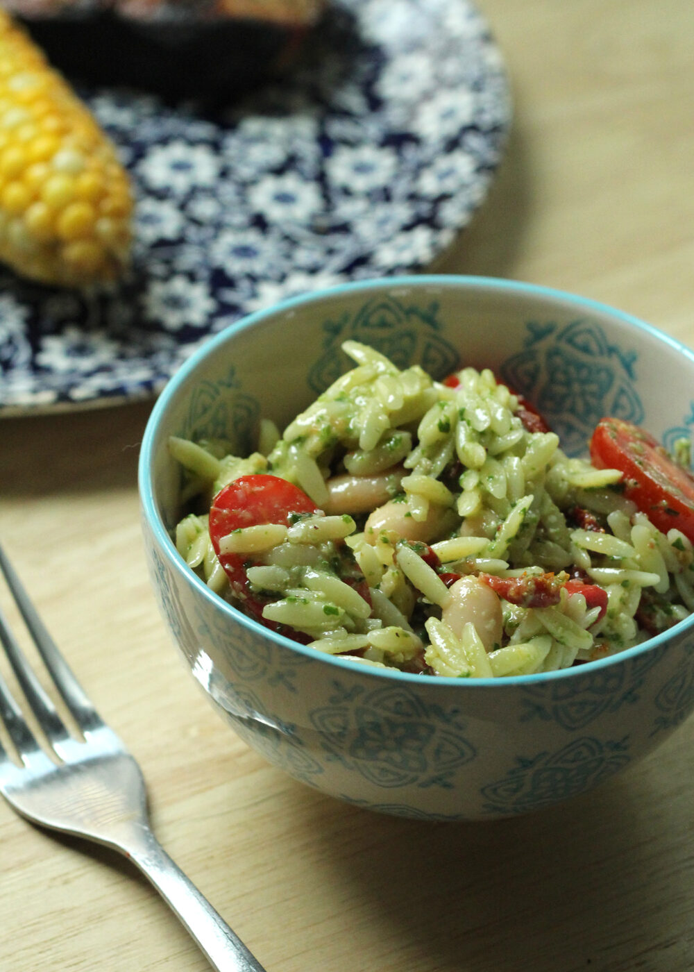 A light blue and white patterned bowl holds oblong orzo, red tomatoes, and creamy colored beans with a green flecked sauce. In the background is a blue and white flowered plate with an ear of corn.