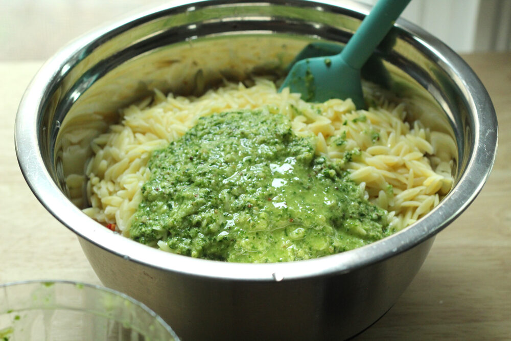 A silver mixing bowl holds creamy colored orzo topped with a green flecked sauce. A blue spatula is stuck in the bowl.