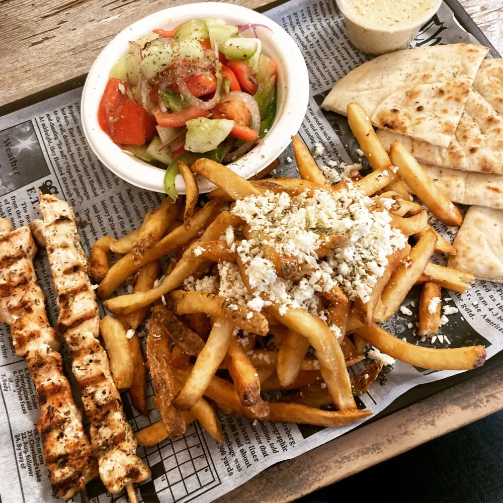 A newspaper lined tray is topped with a pile of fries with feta and herbs as well as chicken skewers, pita bread, tan hummus and a red tomato and green cucumber salad with red onions.