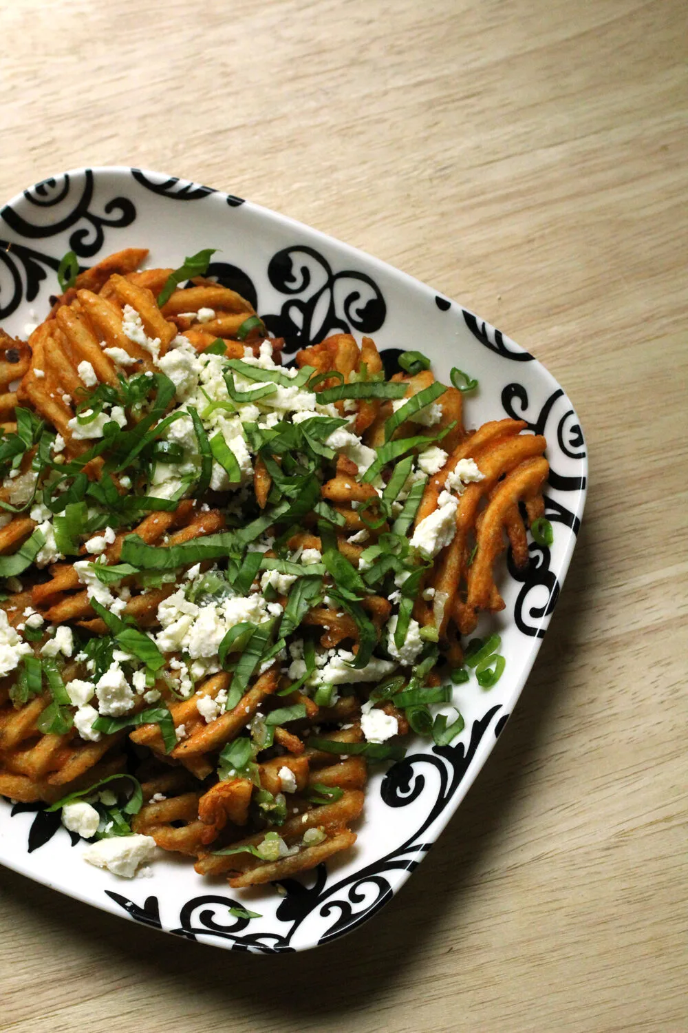 A black and white plate is filled with waffle fries topped with white feta cheese and green basil.