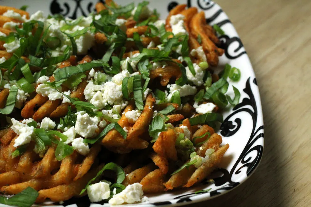 A black and white plate is filled with waffle fries topped with white feta cheese and green basil.