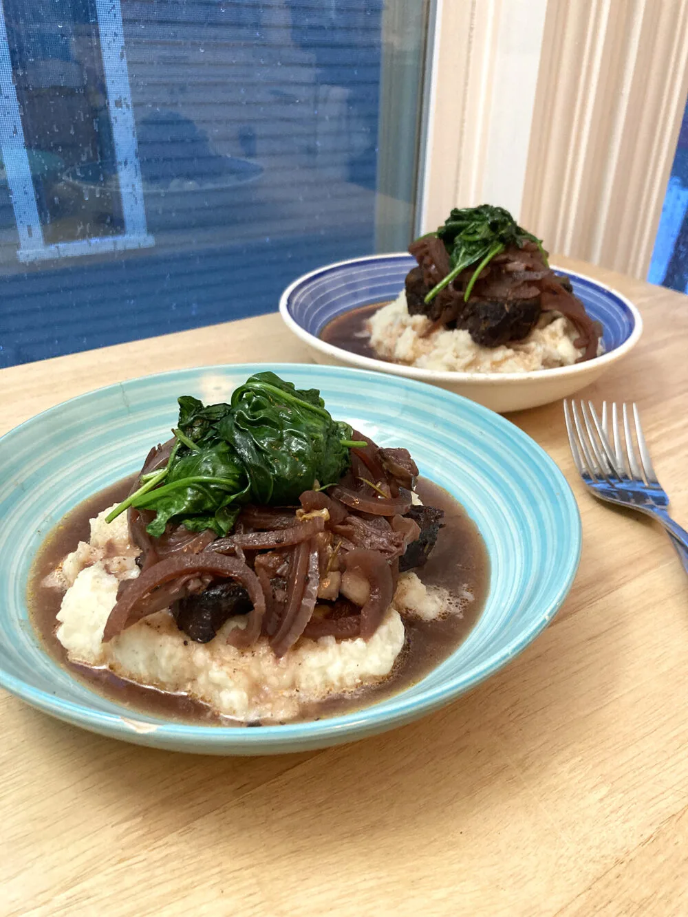 Two shallow bowls are shown on a wooden countertop. Each has a pile of mashed potatoes topped with brown short ribs and onions and green sauteed spinach. 