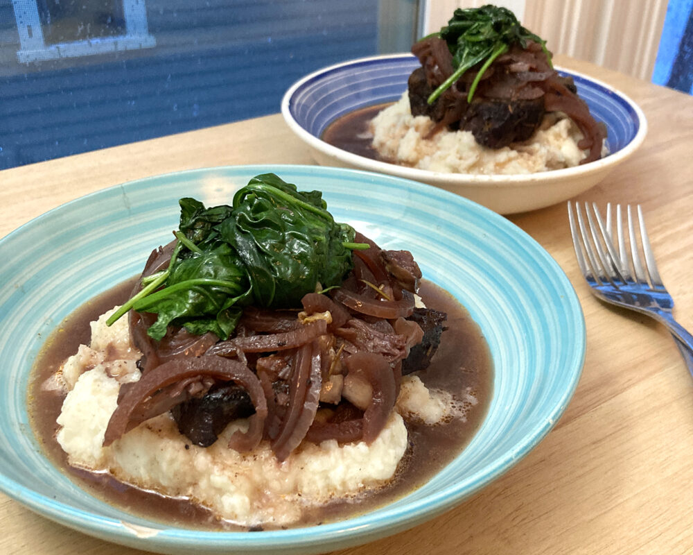 Two shallow bowls are shown on a wooden countertop. Each has a pile of mashed potatoes topped with brown short ribs and onions and green sauteed spinach. 