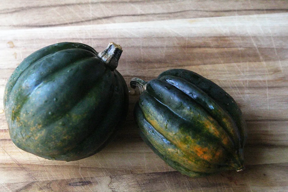 Two acorn squashes are shown on a wooden cutting board. They have a pointy end and lengthwise groves and are mostly a deep green in color.