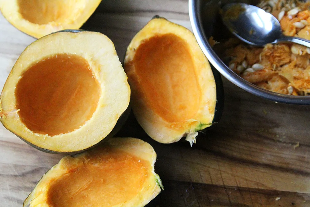 Four halves of acorn squash are shown in a wooden cutting board, cut side up. The flesh is orange. A bowl with seeds and stringy insides is shown nearby.