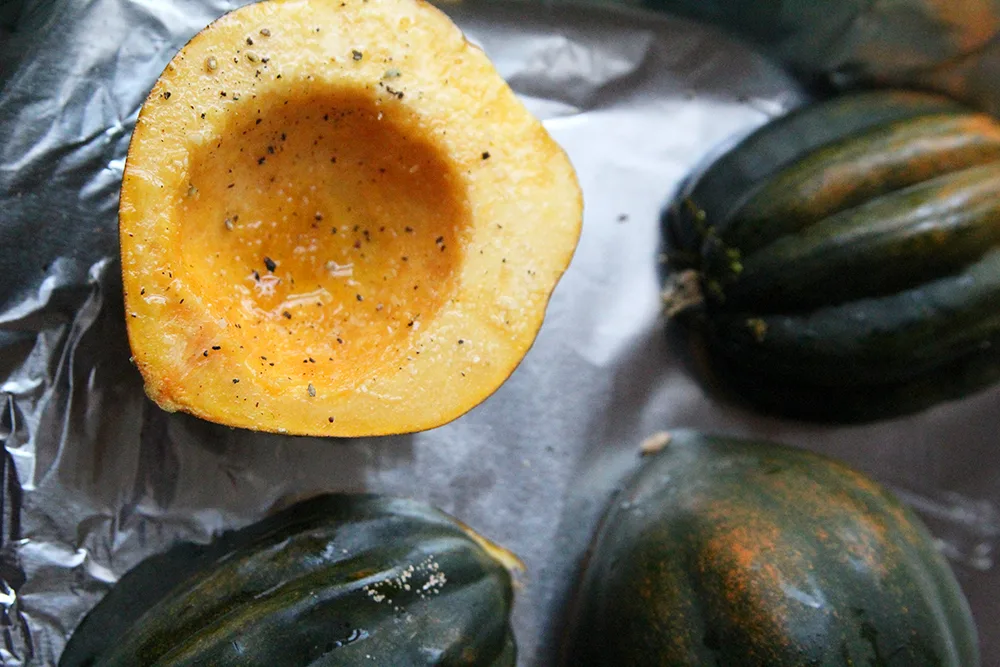 One half of acorn squash is shown on a baking sheet, cut side up. The flesh is orange. Three halves are shown cut-side down. The exterior is mostly rich green.