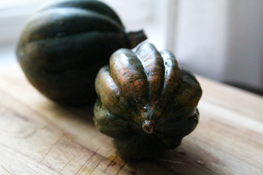 Two acorn squashes are shown on a wooden cutting board. They have a pointy end and lengthwise groves and are mostly a deep green in color.