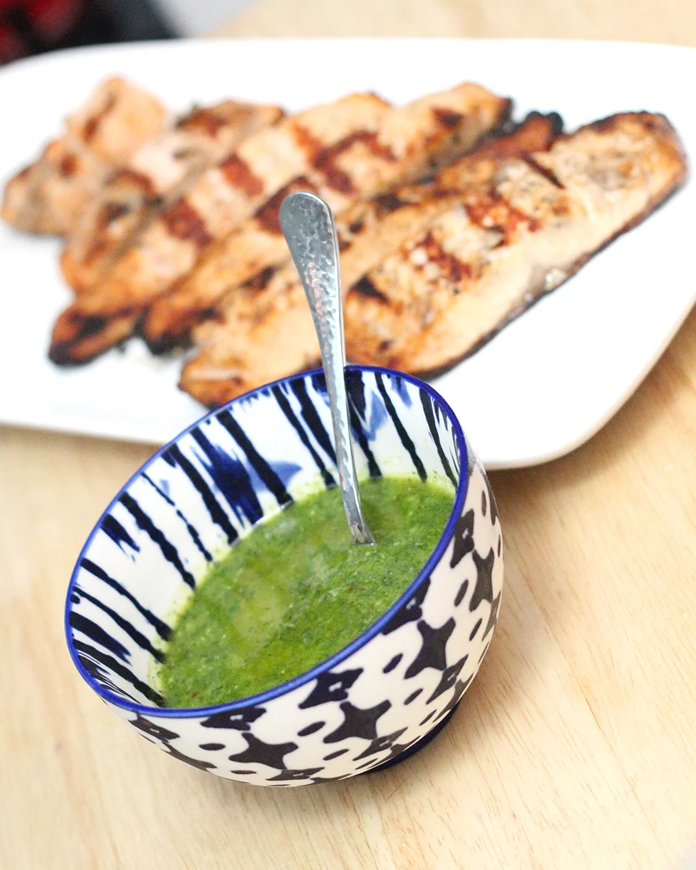 A white platter holds pink-orange fillets with grill marks. A blue and white bowl holding a green sauce and a silver spoon sit nearby.