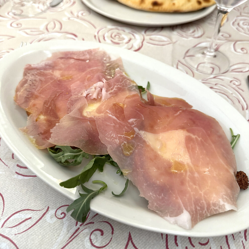 On a white plate sits a an open face sandwich on top of green arugula with a touch of olive oil. The bread topped with barely visible white burrata cheese and draped with a pink-orange slice of prosciutto with white veins of fat.