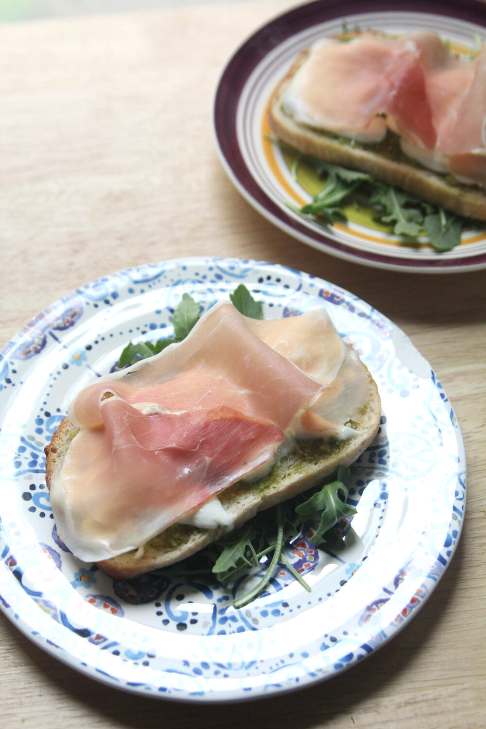 On a white plate with a blue and purple pattern sits a slice of bread on top of green arugula. The bread is spread with green pesto, topped with white burrata cheese and draped with a pink-orange slice of prosciutto with white veins of fat.