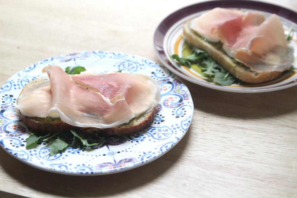On a white plate with a blue and purple pattern sits a slice of bread on top of green arugula. The bread is spread with green pesto, topped with white burrata cheese and draped with a pink-orange slice of prosciutto with white veins of fat.