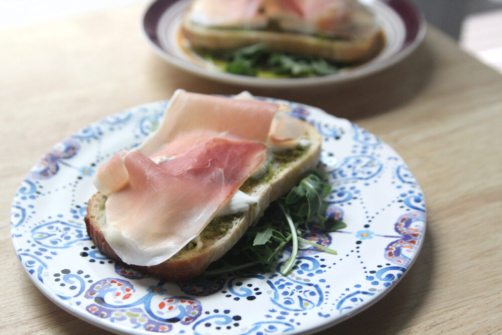 On a white plate with a blue and purple pattern sits a slice of bread on top of green arugula. The bread is spread with green pesto, topped with white burrata cheese and draped with a pink-orange slice of prosciutto with white veins of fat.