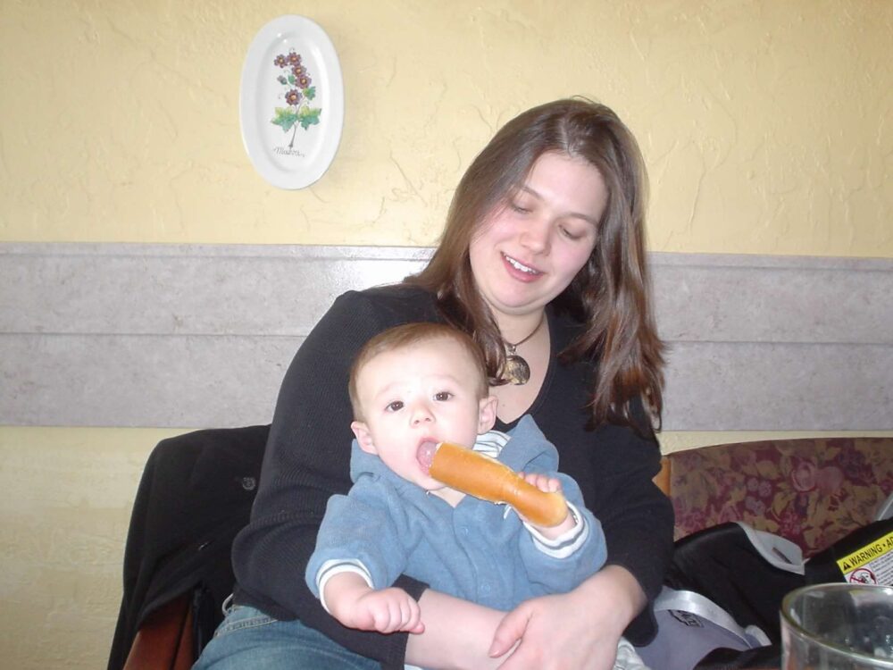 A woman smiles at a baby she's holding while the baby bites a soft breadstick.