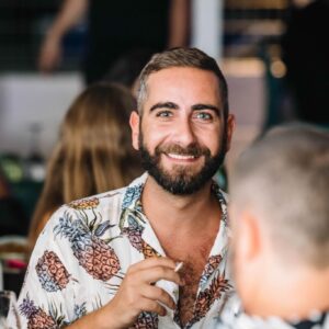 A smiling man with brown hair and a beard looks at the camera. He's wearing a button down shirt with pineapples.