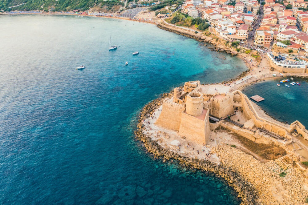 An aerial shot of Calabria in Italy is shown. There is rich blue water, a coastline and tan buildings. Some have red roofs.